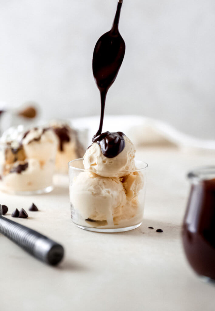 A spoon drizzling hot fudge on vanilla ice cream that is scopped in a glass 