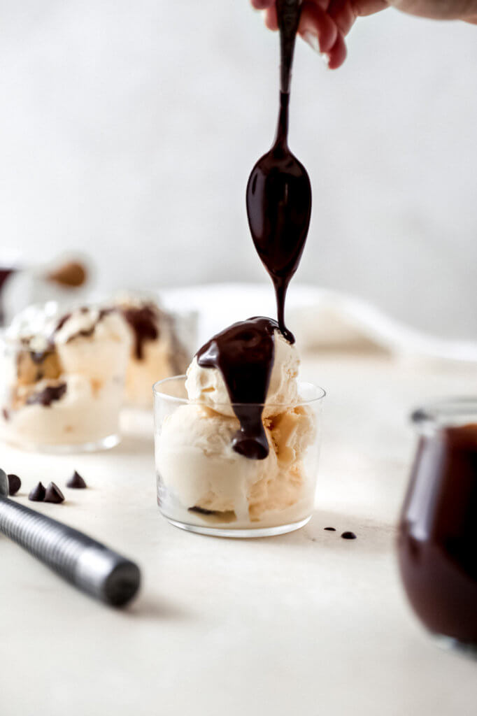 A glass filled with vanilla ice cream with a spoon drizzling dairy-free hot fudge on top. Chocolate chips and ice cream scoops scattered around. 